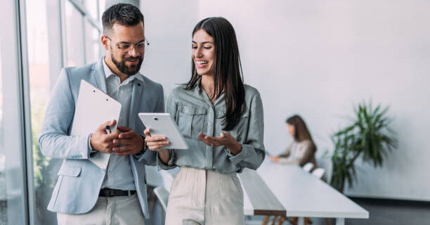 Shot of a two confident business persons talking in the work place. Two colleagues using a digital tablet while walking in a modern office. Businessman and businesswoman in meeting discussing business strategy. Business coworkers working together in the offic